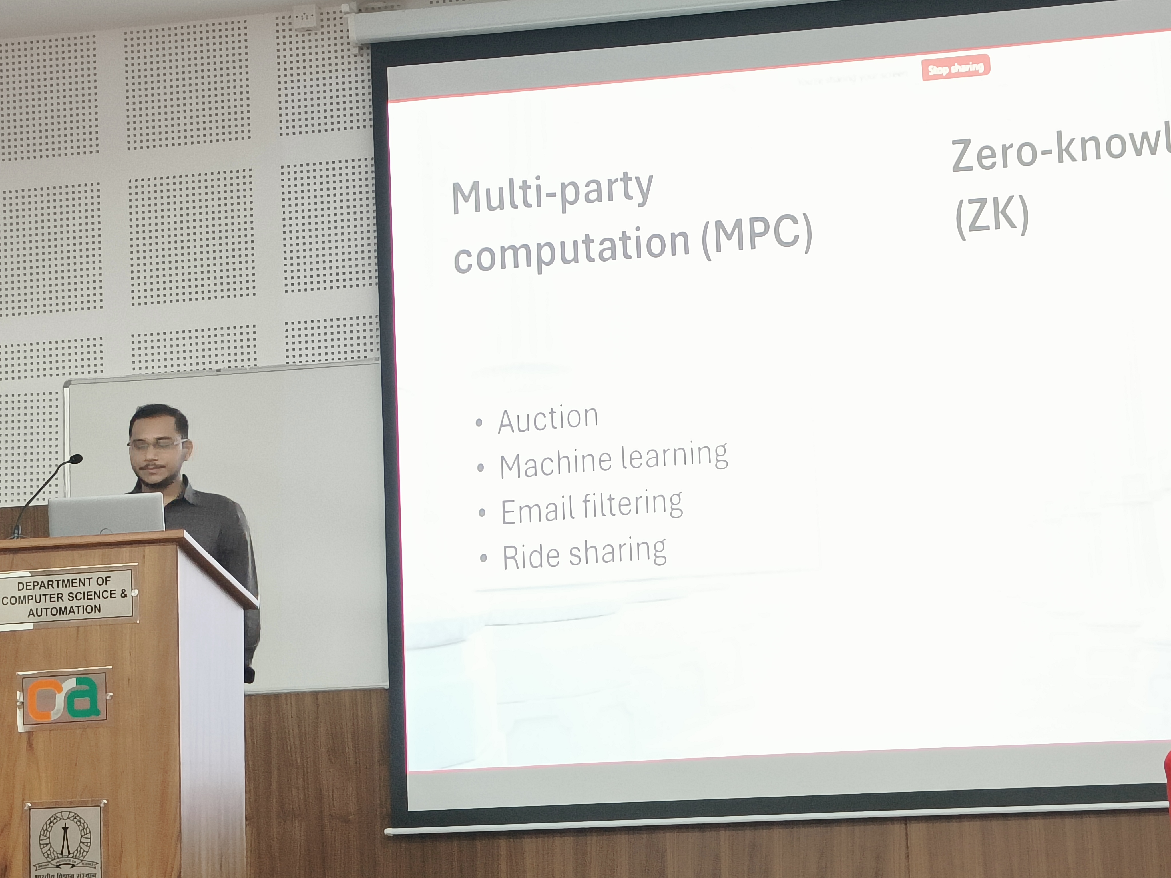 Protik presenting at the podium, with a slide on multi-party computation and zero-knowledge